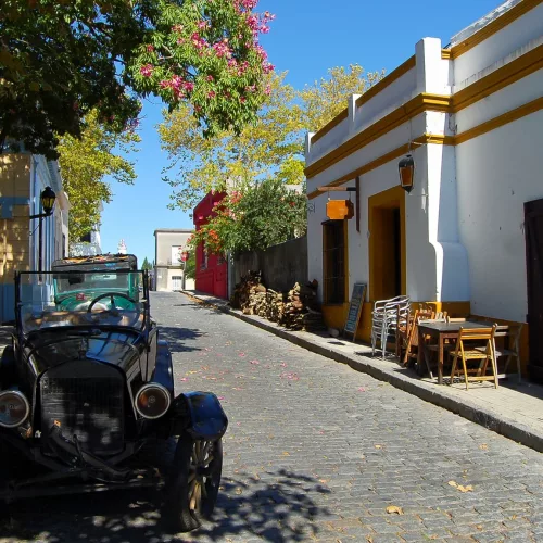 Rua de Colonia do Sacramento, no Uruguai, com mesinhas do lado de fora de prédios e um carro antigo estacionado embaixo de uma árvore