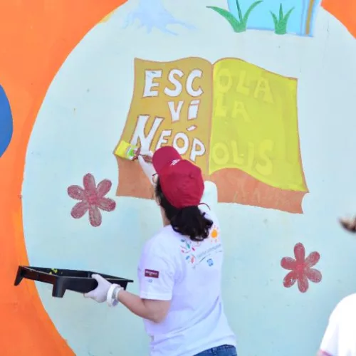 Atividade Colorful Communities na escola. Uma mulher pinta um muro onde se vê a imagem de um livro e o nome escola Vila Neópolis