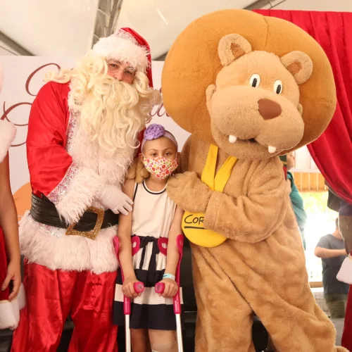 Instituto do Câncer Infantil. Uma pequena paciente, usando máscara e muletas, entre o Papai Noel e o leão-mascote do ICI. Nas pontas, ajudantes de Papai Noel.