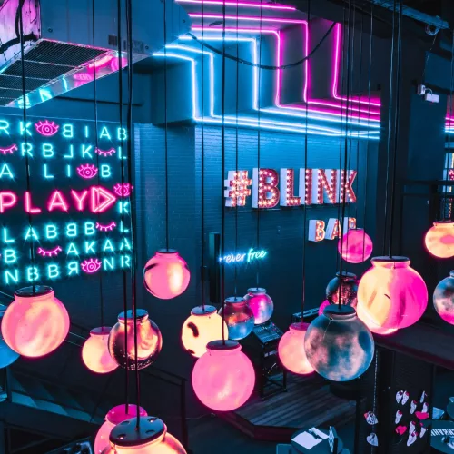 Blink Bar. Decoração em azul, luminárias em rosa e salmão iluminadas, na parede lê-se #Blink