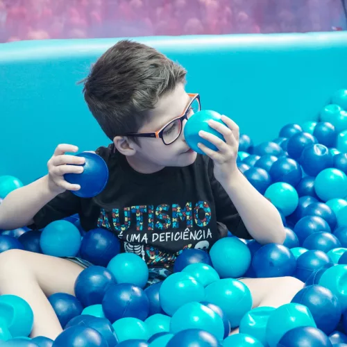 Diversão inclusiva. Um menino brinca com bolinhas em piscina cheia delas. Na camiseta que ele veste pode ser lida a palavra autismo.
