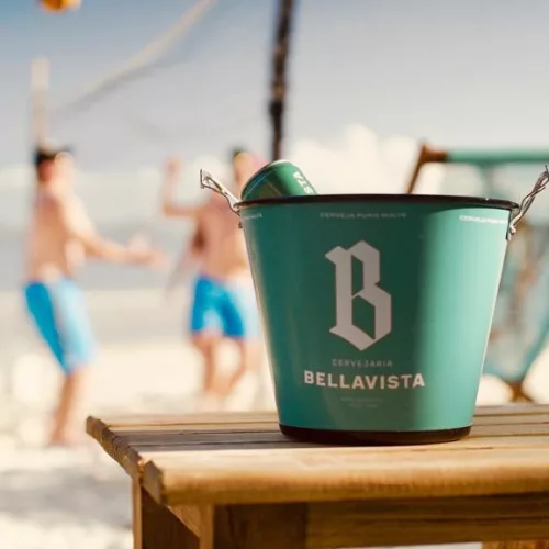 Encontros reais. Em primeiro plano um balde com latinha de cerveja e ao fundo pessoas interagem em uma praia.