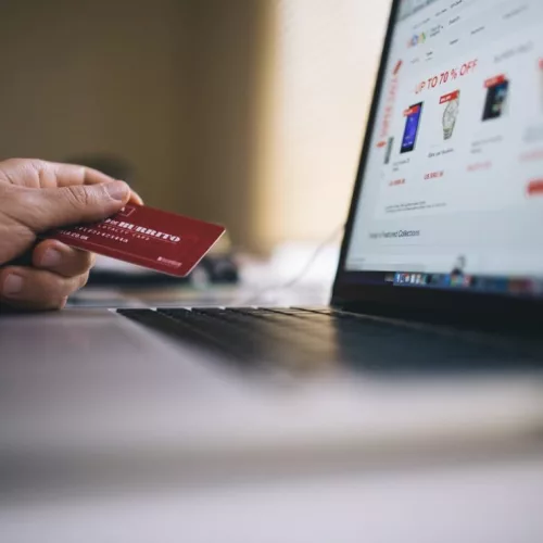 E-commerce. Uma mão segura um cartão de crédito em frente a um notebook aberto em um e-commerce.