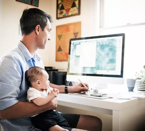 Oi Soluções. Um homem segura um bebê enquanto trabalha em frente ao computador.