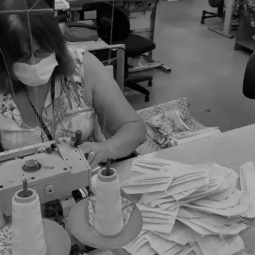 AMC Têxtil. Mulher com máscara produz mais máscaras em máquina de costura. Foto em P&B.