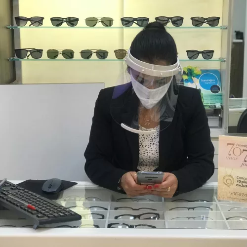 Safira. Mulher manuseia celular em cima de balcão de vidro de loja de joias. Ela usa máscara e escudo de proteção.