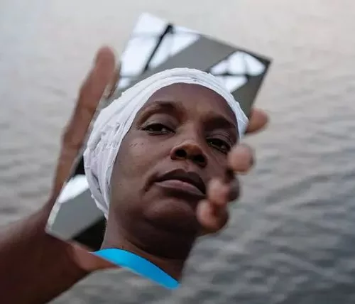 O reflexo de uma mulher negra em um espelho que uma pessoa segura na mão. Ao fundo, o mar.