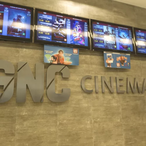 GNC Cinemas escrito na parede e acima tvs com a programação.