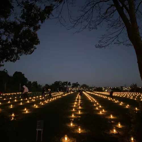 Várias velas acesas à noite em um campo com as pessoas circulando por ele.