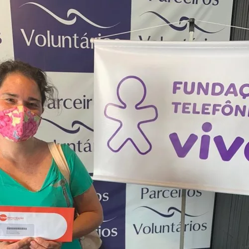 Mulher segura envelope ao lado de uma faixa da Fundação Telefônica Vivo e em frente a um banner da ong Parceiros Voluntários.
