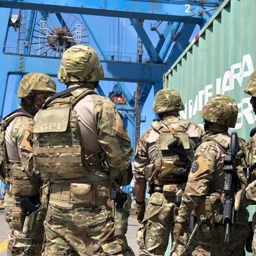 Militares participam de simulação de segurança portuária. Homens vestidos com roupas do exército em um porto num dia de céu azul.