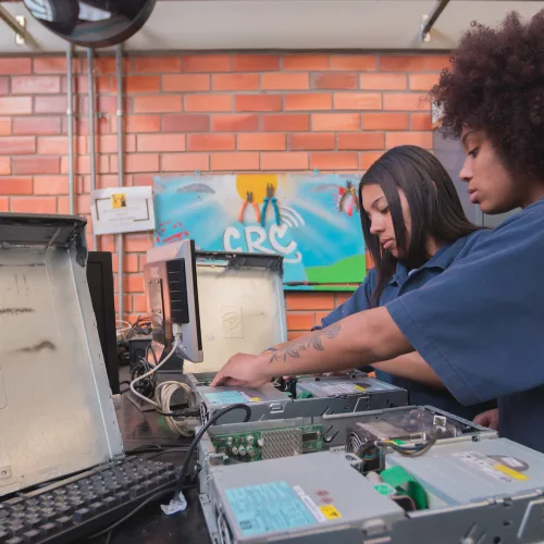 Meninas trabalham em computadores abertos. Ambas negras, a da frente com cabelo black power, a de trás com cabelo liso.