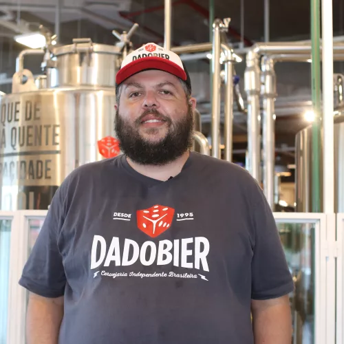 Um homem de barba usa boné e camiseta Dado Bier em frente à produção de cerveja.
