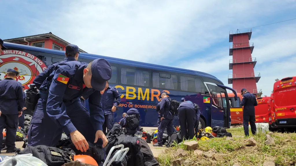 Bombeiro militar está agachado se preparando para embarcar para o Taim. Ele veste roupa azul e boina. Ao fundo, outros militares e um ônibus dos Bombeiros.