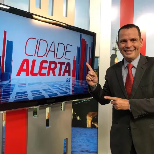 Jornalista Voltaire Porto posa ao lado de um monitor no estúdio da TV Record RS, em Porto Alegre. Ele aponta para a tela, onde se lê "Cidade Alerta RS".