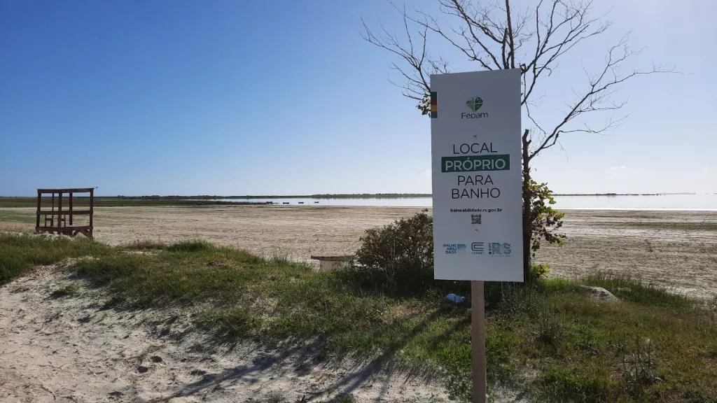 Placa da Fepam com a inscrição "local próprio para banho". Ao fundo, praia vazia e céu azul compõem a paisagem. 