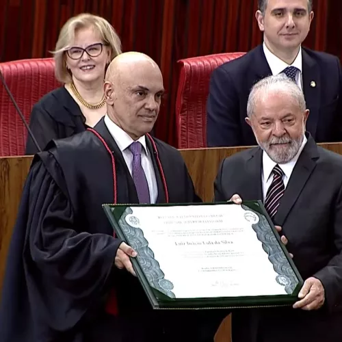 O presidente do TSE, Alexandre de Moraes e o presidente eleito Lula seguram o diploma. Moraes está com uma toga preta. Lula de terno e gravata vermelha.