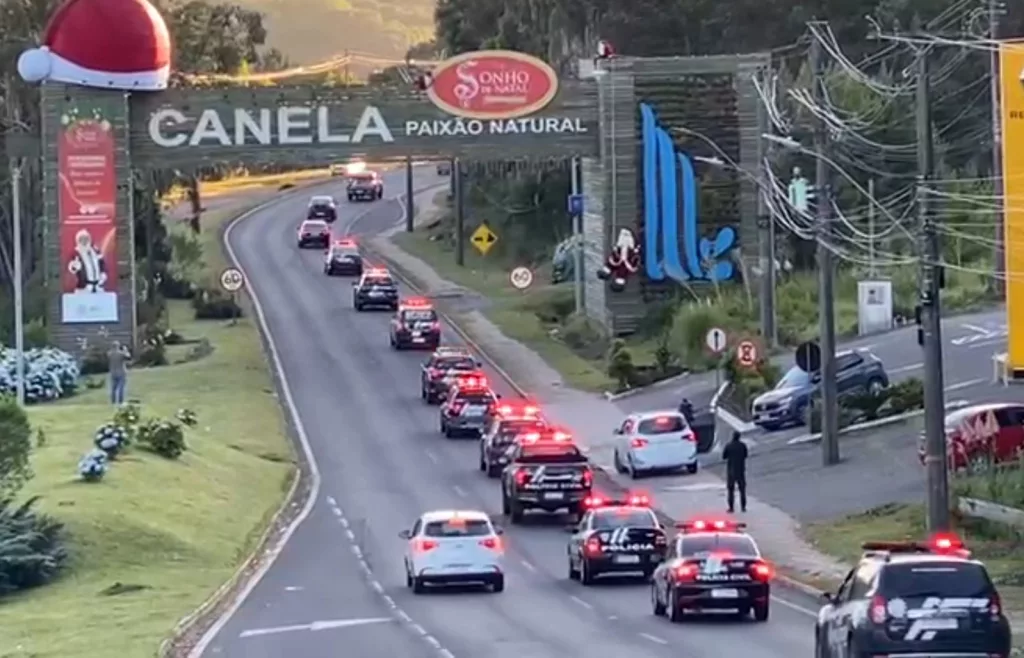 Treze viaturas da Polícia Civil estão em fila, andando em uma estrada. Elas estão passando pelo pórtico de entrada de Canela, na Serra do RS.