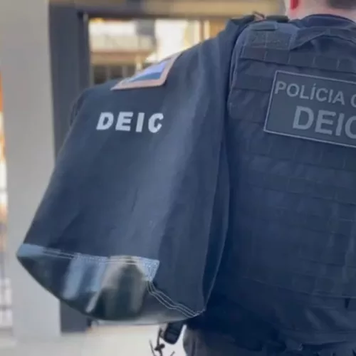 Policial civil veste um colete à prova de balas e carrega um malote com a expressão "DEIC". Ele está de costas e é possível ler "Polícia Civil DEIC". Ao fundo, é possível ver uma grade e um portão residencial.