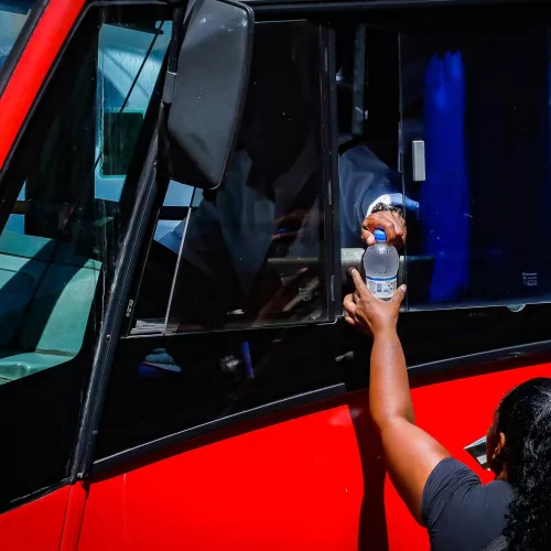 Mulher entrega uma garrafa de água para o motorista de um táxi-lotação.