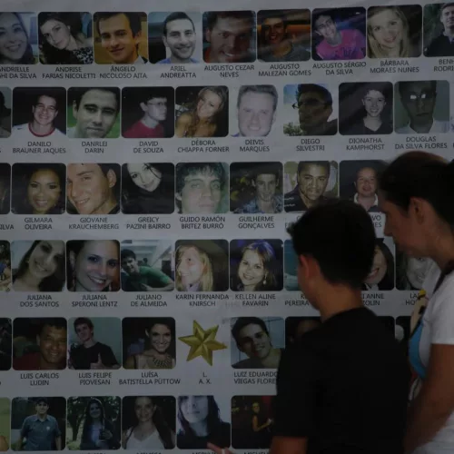 Mulher e menino olham para painel onde estão nomes e fotos das vítimas do incêndio na boate Kiss, em Santa Maria, no ano de 2013.