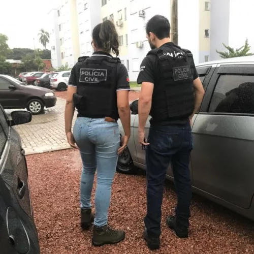 Dois policiais, um homem e uma mulher, de uniforme e de costas para a câmera, durante cumprimento de mandados da operação figueiras