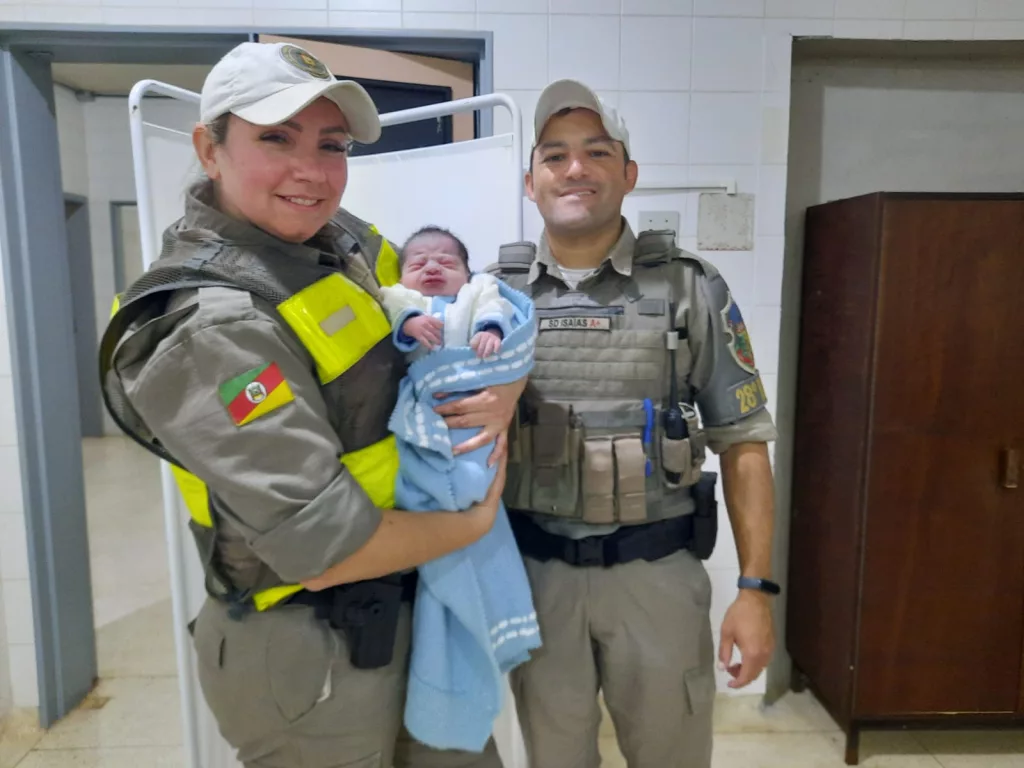 Rodrigo é segurado pela soldado Luciana. Ela é acompanhada pelo soldado Isaías, do 28° BPM. Ambos vestem farda da Brigada Militar e estão posando para a foto em um ambiente hospitalar.