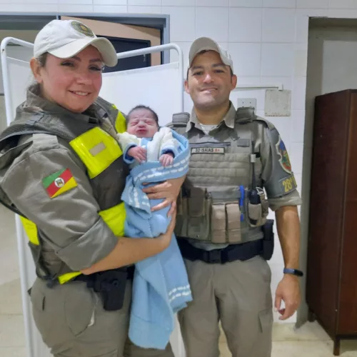 Rodrigo é segurado pela soldado Luciana. Ela é acompanhada pelo soldado Isaías, do 28° BPM. Ambos vestem farda da Brigada Militar e estão posando para a foto em um ambiente hospitalar.