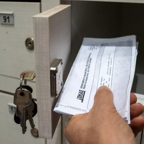 Uma pessoa pega, com a mão esquerda, uma conta do DMAE colocada em uma gaveta de um condomínio residencial.