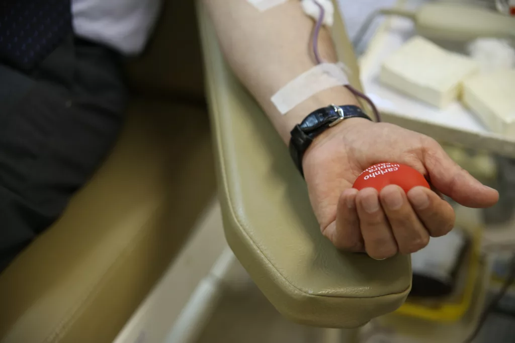 Homem está em uma cadeira, em ambiente hospitalar, com o braço esquerdo estendido enquanto faz doação de sangue. Ele segura uma bolinha vermelha.
