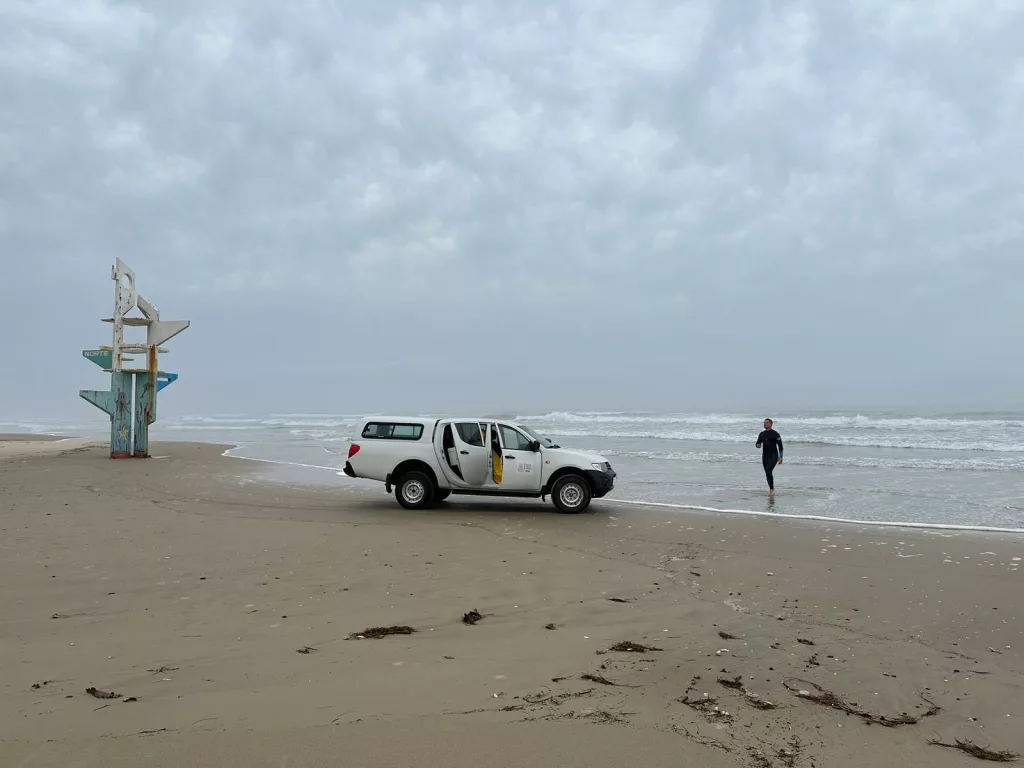 Imagem de um carro da Fepam em uma praia com uma pessoa saindo do mar com a coleta e água para análise do Projeto Balneabilidade 2023-2024.